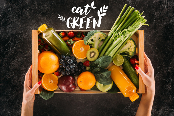 partial view of woman holding wooden box with healthy vegetables, fruits and detox drinks with "eat green" lettering - Foto, Bild