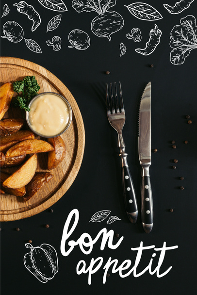 top view of baked potatoes with sauce on wooden board and fork with knife on black with "bon appetit" lettering - Foto, afbeelding