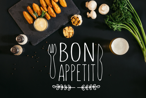 top view of delicious baked potatoes with sauce, spices and glass of beer on black with "bon appetit" lettering with fork and knife - Photo, Image