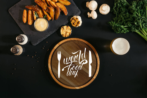 wooden board  with "world food day" lettering with fork and knife, glass of beer and baked potatoes with sauce and spices on black - Photo, Image