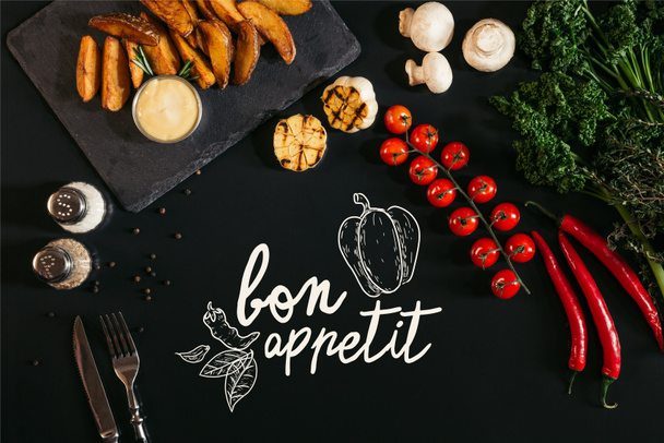 top view of tasty baked potatoes with sauce, spices and vegetables on black with "bon appetit" lettering - Fotografie, Obrázek