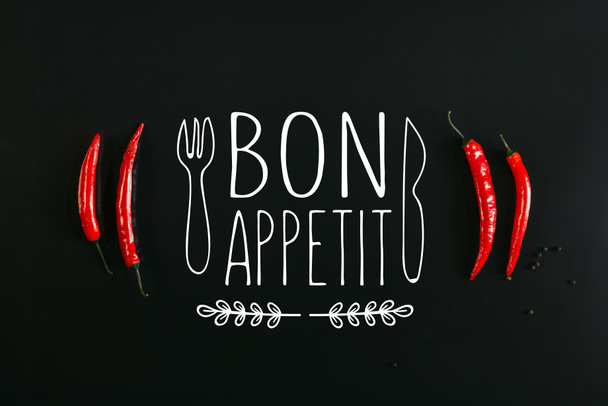 top view of red chili peppers and peppercorns on black background with "bon appetit" lettering with fork and knife - Photo, Image