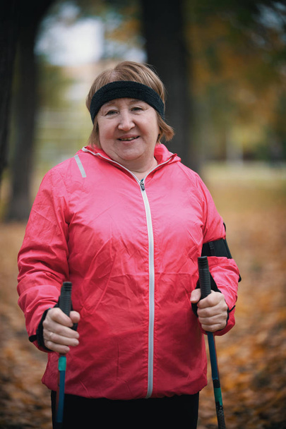 Eine ältere Frau in pinkfarbener Daunenjacke steht mit Stöcken für Nordic Walking im Park - Foto, Bild