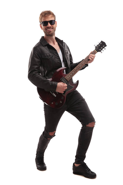 joven estrella del rock en chaqueta de cuero actuando en la guitarra y sonriendo mientras está de pie sobre fondo blanco
 - Foto, imagen