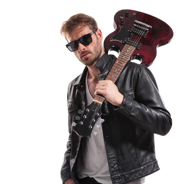 portrait of cool rock star with sunglasses holding his guitar on shoulder while standing on white background - Photo, Image