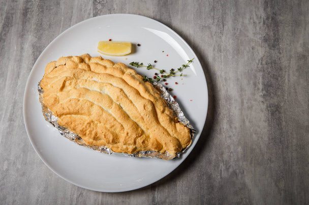 Bovenaanzicht op gebakken dorado onder het opgeklopte eiwit op folie met kruiden en citroen op witte restaurant plaat op grijze tafel geserveerd - Foto, afbeelding