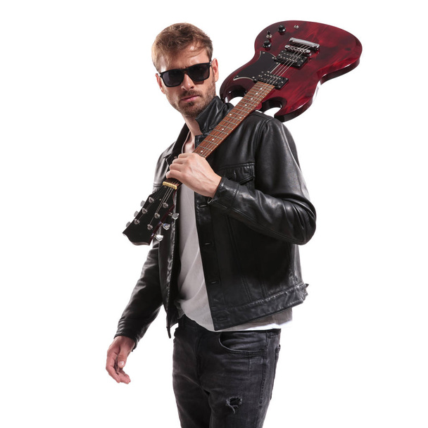 portrait of cool guitarist in black leather jacket posing with his electric guitar on shoulder while standing on white background - Foto, afbeelding