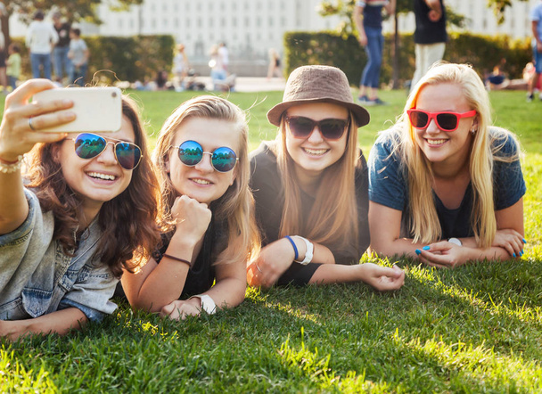 Grupo de mujeres jóvenes y elegantes felices sonrientes y despreocupadas hacen amigos selfie a su teléfono inteligente acostado en la hierba en el parque, concepto milenario, jóvenes y estudiantes, turismo y vacaciones
 - Foto, imagen