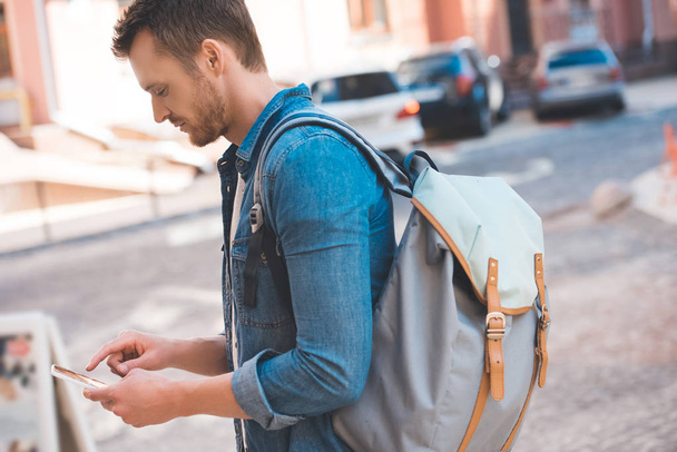 handsome young man with backpack walking by street and using smartphone - Foto, Bild