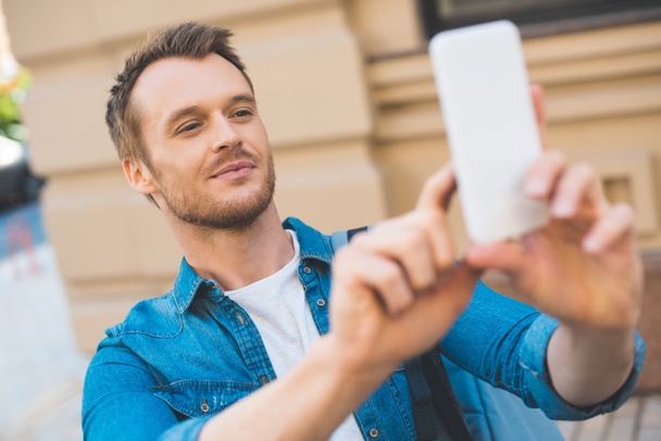 close-up portrait of handsome young tourist with backpack taking photo with smartphone on street - Photo, Image