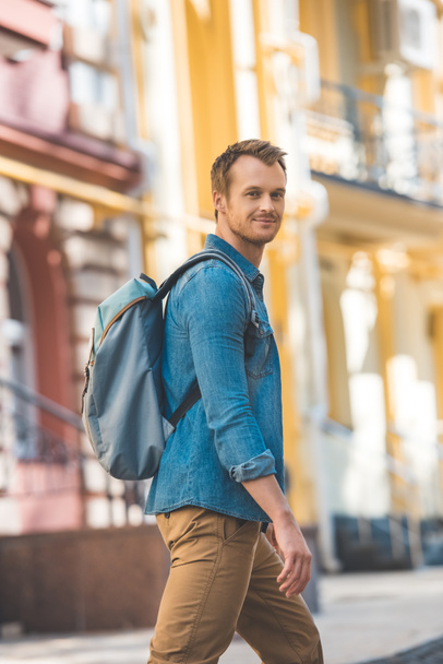 attractive young traveller with backpack walking by street and looking at camera - Foto, Imagem