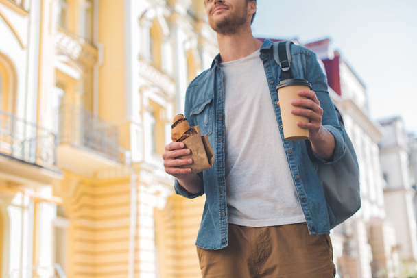 cropped shot of happy man with backpack, coffee to go and croissant walking by street - Foto, imagen