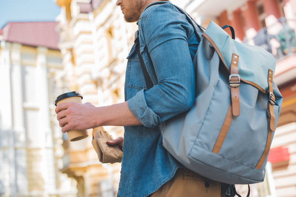 plan recadré de jeune homme avec sac à dos, café à emporter et croissant marchant dans la rue
 - Photo, image
