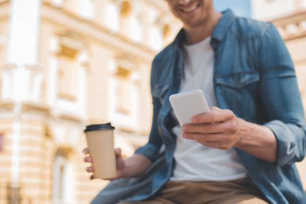 cropped shot of smiling young man with coffee to go using smartphone on street - Φωτογραφία, εικόνα
