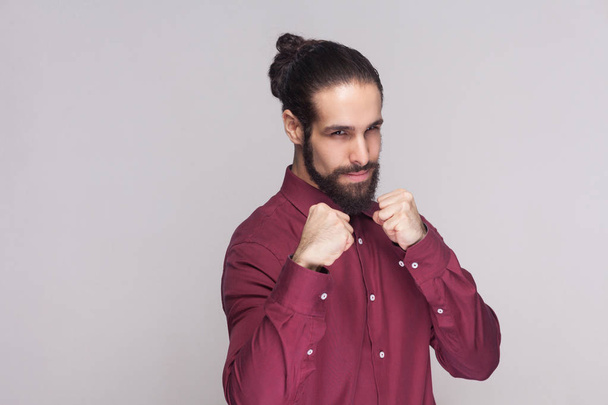 Portrait of serious handsome man with dark collected long hair and beard in red shirt standing with boxing fists and ready to attack on gray background - Foto, Imagen