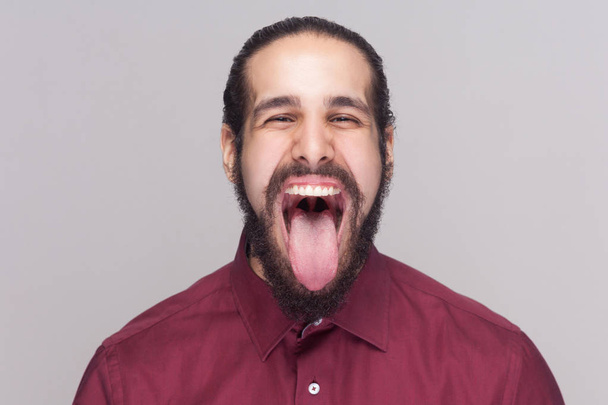 Vicces crazy jóképű férfi szakállal látszó-on fényképezőgép nagy nyitott száj és nyelv, és sikoltozik a szürke háttér piros inget portréja - Fotó, kép