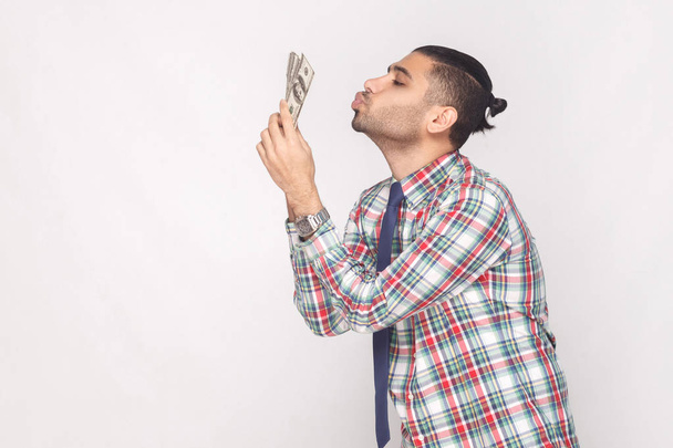 side view of cheerful rich handsome bearded young stylish businessman in colorful checkered shirt air kissing fan of dollars on grey background - 写真・画像
