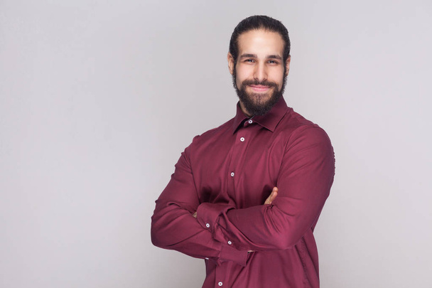 Portrait of handsome businessman with dark hair and beard in red shirt standing with crossed hands and looking at camera with satisfied face on gray background - Photo, image
