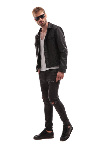 side view of young blond man wearing sunglasses and leather jacket stepping to side on white background, full body picture - Photo, Image