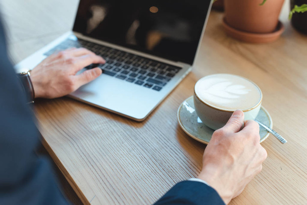 bijgesneden schot van zakenman met kopje koffie werkt op laptop met leeg scherm in café - Foto, afbeelding