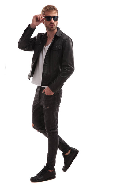 relaxed handsome man in black leather jacket steps to side on white background and fixes sunglasses, full length picture - Fotó, kép
