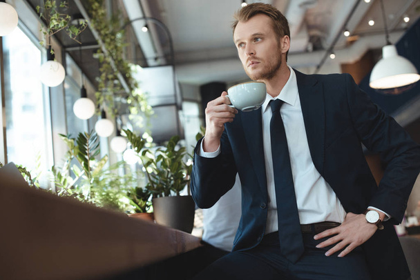 stylish thoughtful businessman in suit with cup of coffee having coffee break in cafe - Foto, Bild