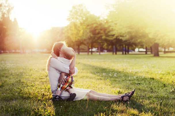 Happy toddler boy relaxing on green grass with mother in sunny park - 写真・画像