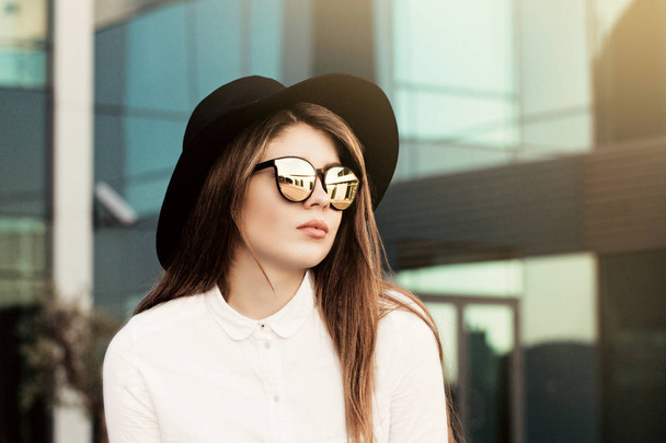 Außenporträt eines hübschen Mädchens mit trendiger Sonnenbrille mit schwarzem Hut und weißem Hemd - Foto, Bild