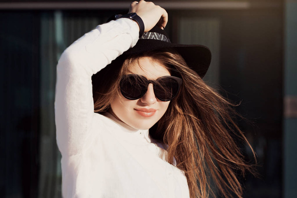 Egy csinos lány a divatos napszemüveg, fekete kalapot és fehér inget viselő szabadtéri portréja - Fotó, kép