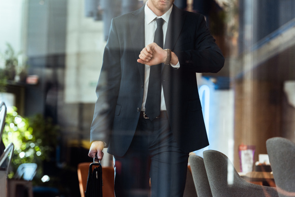 partial view of businessman with suitcase checking time while walking in cafe - Foto, Imagem