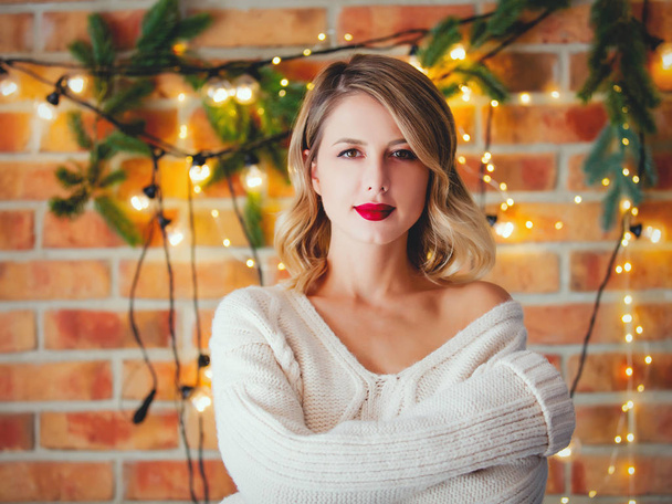 Portrait of a young cozy woman in white sweater with Christmas lights and pine branch on background. - Foto, immagini