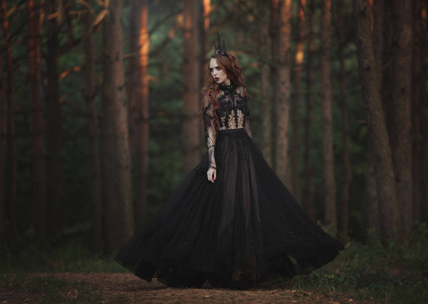 A beautiful gothic princess with pale skin and very long red hair in a black crown and a black long dress in a misty fairy forest. The costume of the dark queen. Artistic processing. - Foto, Imagem