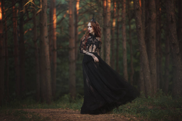 A beautiful gothic princess with pale skin and very long red hair in a black crown and a black long dress in a misty fairy forest. The costume of the dark queen. Artistic processing. - Photo, image
