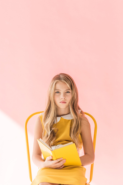 fashionable youngster reading book while sitting on yellow chair on pink - Foto, imagen