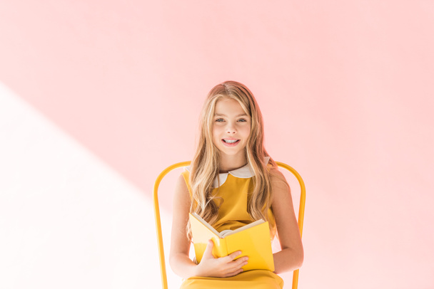 smiling kid reading book while sitting on yellow chair on pink - Φωτογραφία, εικόνα