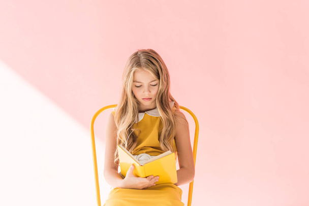 beautiful blonde youngster reading book while sitting on yellow chair on pink - Foto, afbeelding