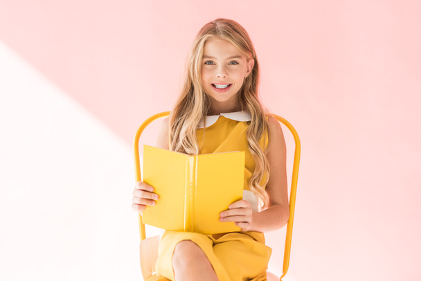 smiling fashionable kid reading book while sitting on yellow chair on pink - Foto, imagen