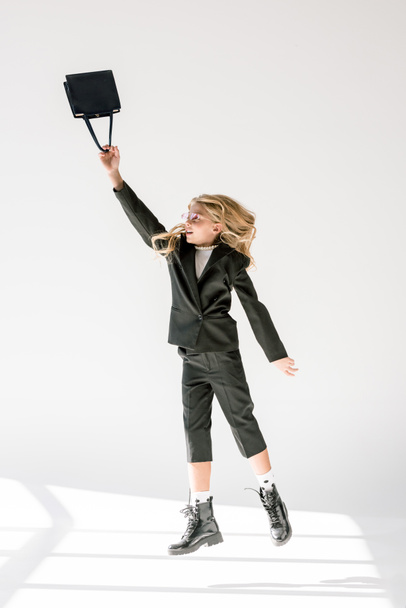 fashionable preteen kid in black suit and pink sunglasses jumping with bag on grey - Fotó, kép
