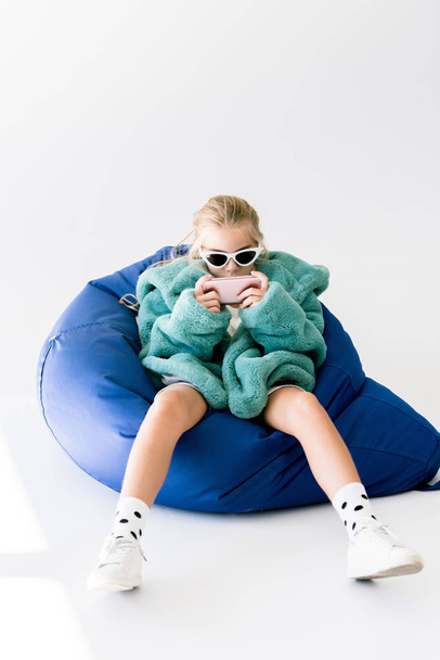 fashionable kid in fur coat and sunglasses using smartphone while sitting in bean bag chair on white - Photo, Image