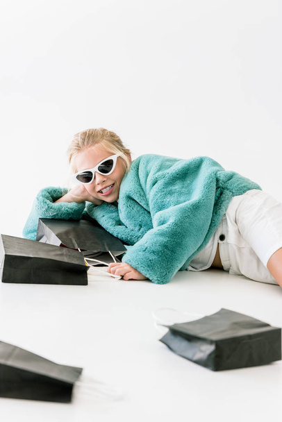 smiling fashionable kid in turquoise fur coat and sunglasses lying with black shopping bags on white - Foto, afbeelding