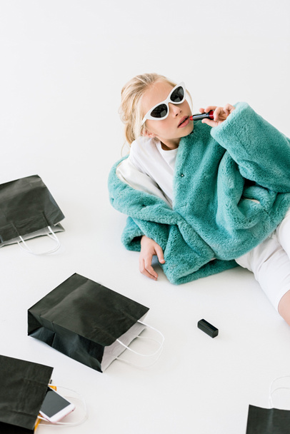 stylish child applying lipstick while lying with black shopping bag on white - Foto, Bild