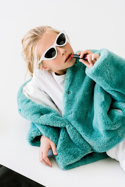fashionable blonde child in turquoise fur coat and sunglasses applying lipstick on white - Foto, afbeelding