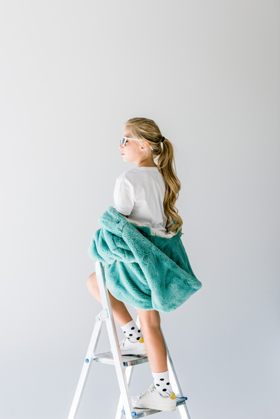 adorable fashionable kid posing in turquoise fur coat on ladder isolated on grey - Fotografie, Obrázek