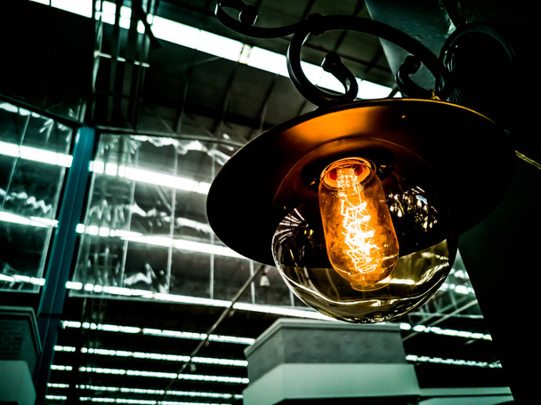 Вольфрамова помаранчева лампа, що звисає всередині будівлі з чорним залізним парканом навколо, Освітлення в будівлях з різнокольоровими електричними лампами, м'який фокус. формат високої контрастності
. - Фото, зображення