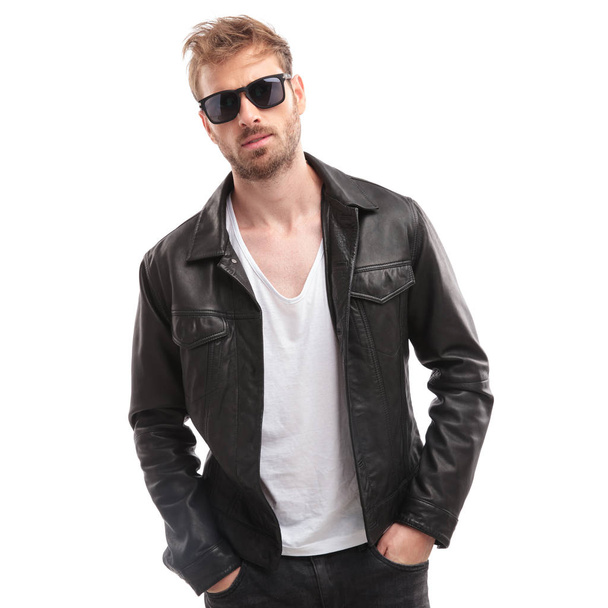 портрет расслабленного мужчины в черных солнцезащитных очках и кожаной куртке на белом фоне с руками в карманах
 - Фото, изображение