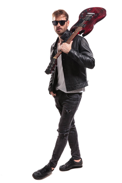 guitarrista relaxado com óculos de sol e jaqueta de couro pisando para o lado enquanto segura guitarra no ombro no fundo branco
 - Foto, Imagem