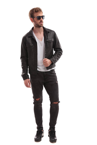 handsome cool man wearing leather jacket and sunglasses looks to side while standing on white background, full length picture - Fotografie, Obrázek