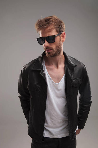 portrait of fashion man wearing sunglasses and leather jacket looking down to side while standing on grey background - Fotografie, Obrázek