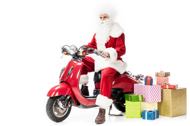 confident santa claus in costume sitting on scooter near gift boxes isolated on white background  - Zdjęcie, obraz