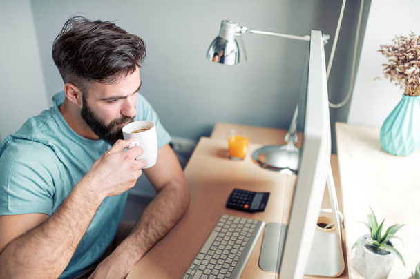 Guapo joven bebiendo café mientras trabaja con el ordenador portátil en casa
 - Foto, Imagen